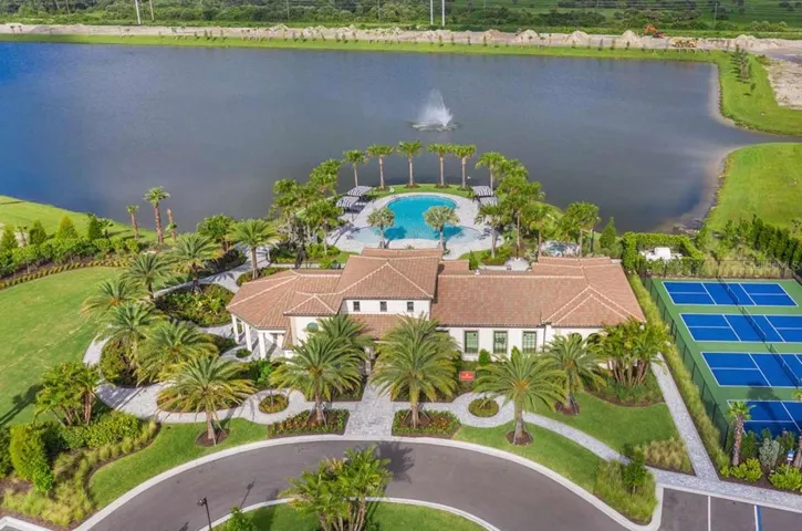An aerial view frames Aria's clubhouse alongside a curving lake and its working fountain, with lighted tennis and pickleball courts, a resort-style pool, and palm-lined walkways below. The scale of the grounds offers a first look at the everyday resort feel awaiting residents just beyond their own front door.