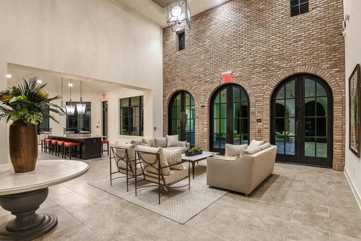 An arched, brick-columned loggia opens onto a shaded outdoor seating area, furnished for residents to relax or socialize before or after using the clubhouse. Wood-beamed ceilings and pendant lighting carry the same finishes found throughout the clubhouse into this open-air gathering spot.