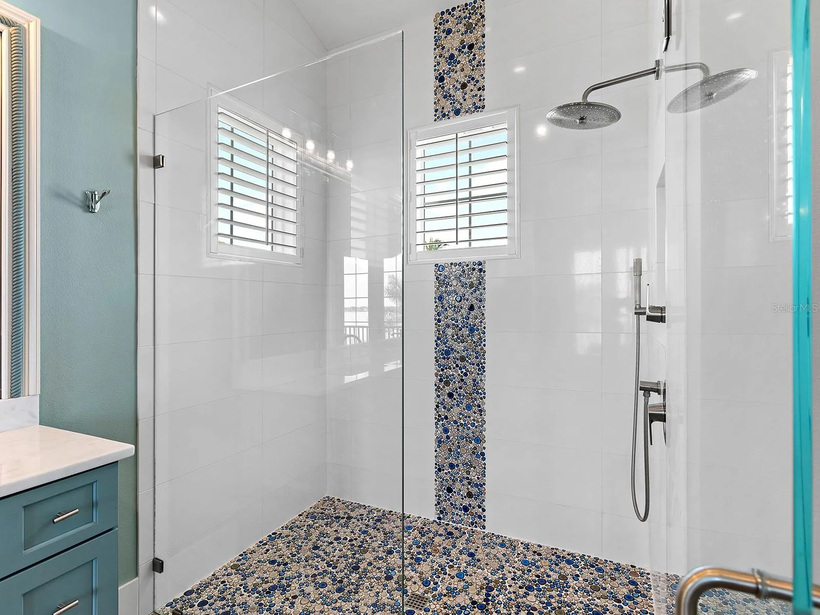 Luxe walk-in shower oasis with a vibrant blue and tan river rock mosaic feature wall, matching pebbled floor, a wall-mounted showerhead, and privacy-enhancing plantation shutters
