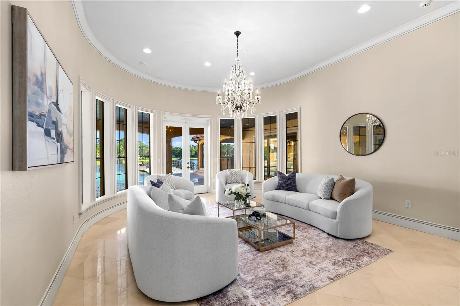 The formal living room is enveloped by a graceful curved wall of windows, creating a luminous, sophisticated setting beneath an elegant crystal chandelier.