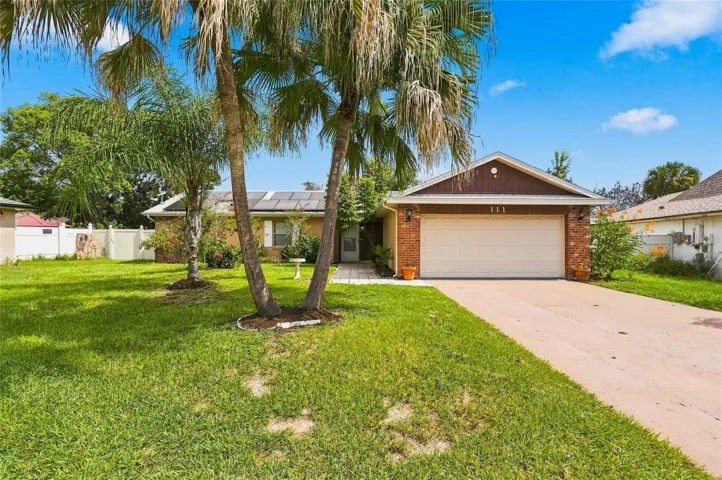 Brick facade residence with a two-car garage, mature palm trees, solar panels, and a concrete driveway