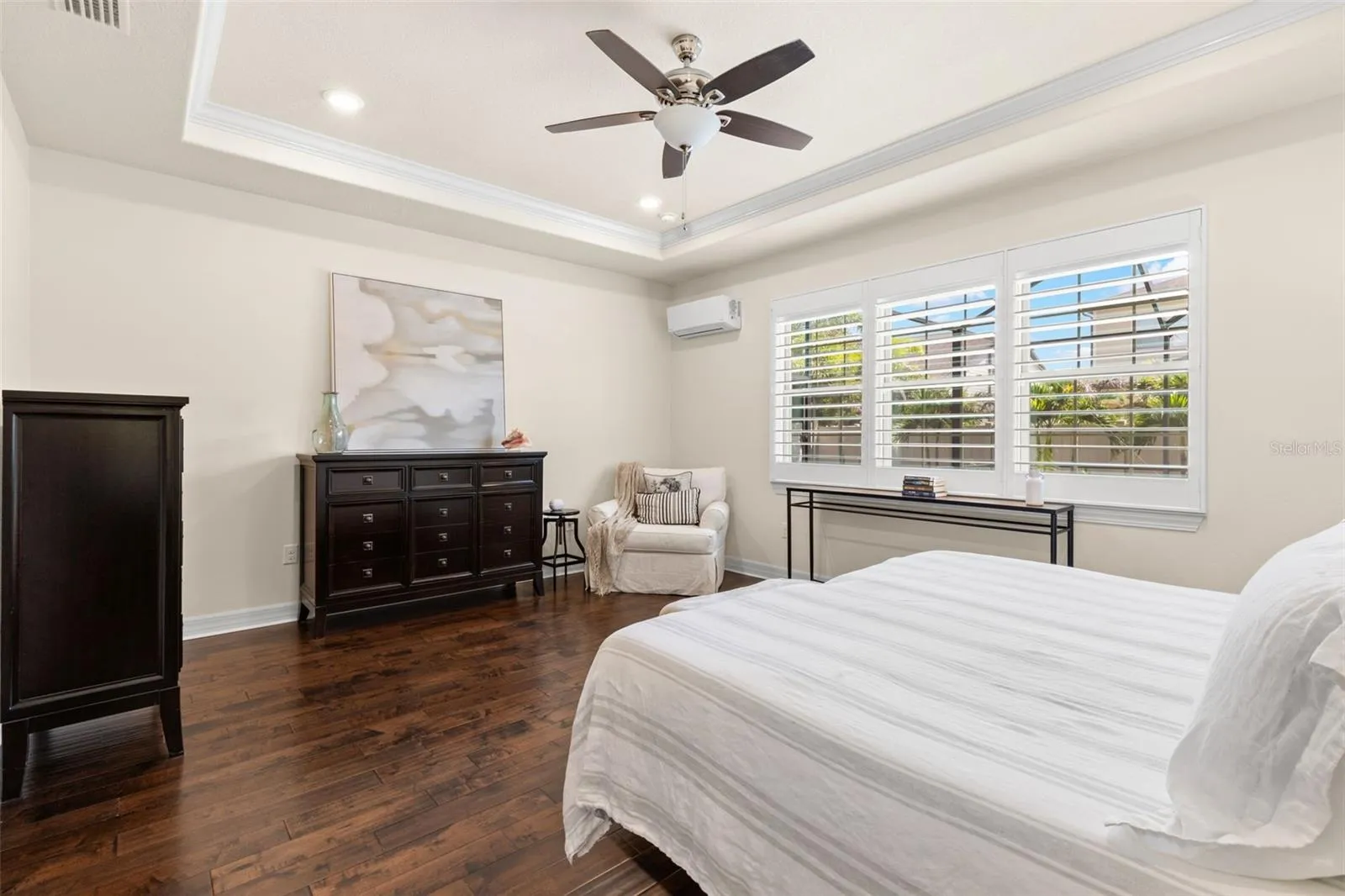 A warm, neutral palette gives the primary bedroom a calm, polished feel, with soft cream walls and crisp trim set against rich, dark engineered hardwood floors. Plantation shutters, a tray ceiling with crown molding, and a ceiling fan complete the comfortably elegant space.