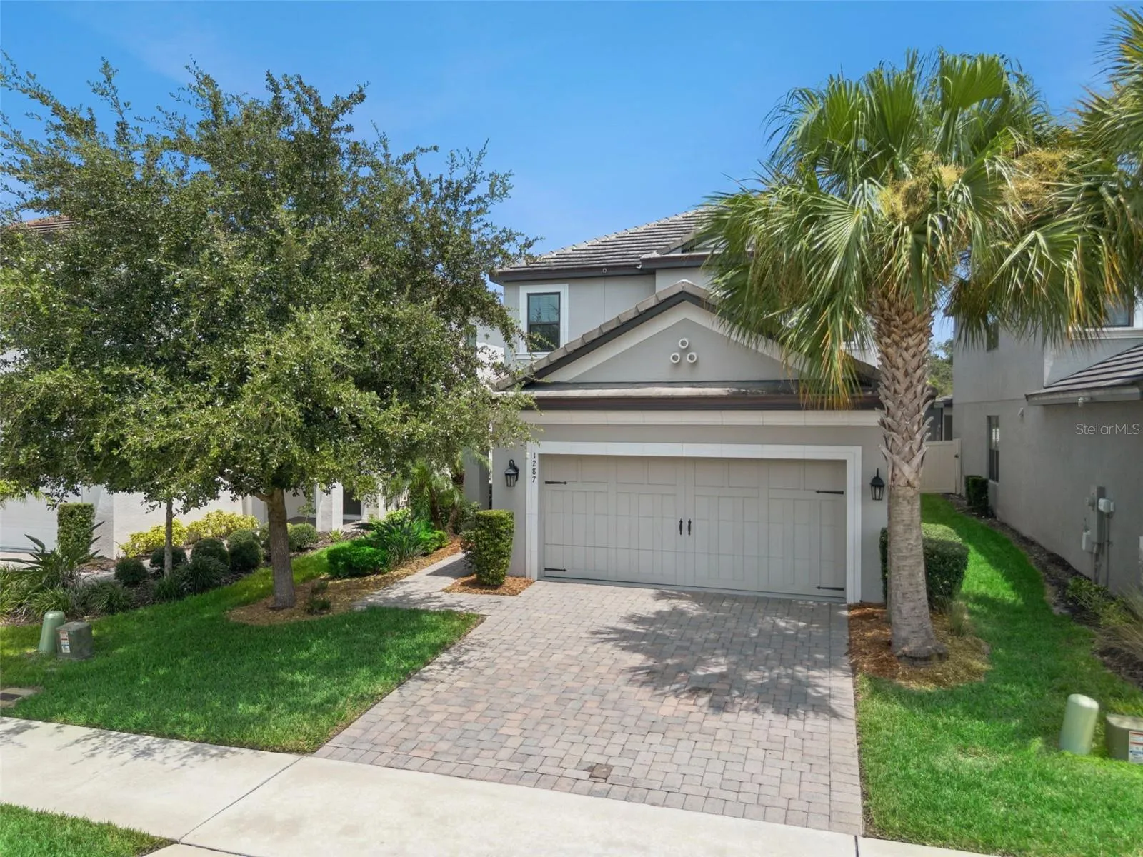 Wide exterior view highlights the two-story design, tile roof, established landscaping, palm trees, and attractive paver driveway.