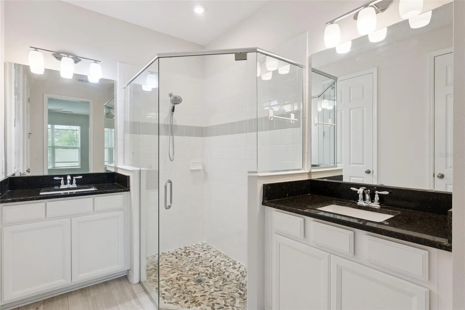 Downstairs Primary Bathroom – Bright ensuite bath features dual vanities with dark stone countertops, abundant white cabinetry, oversized mirrors, and a glass-enclosed tiled shower with a decorative pebble-style floor.