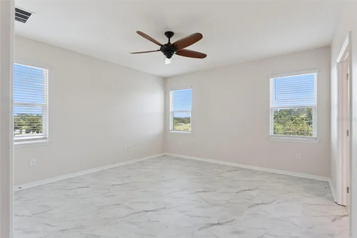 2nd Primary Suite (Upstairs)  – Spacious upstairs primary bedroom with marble-look tile flooring, a ceiling fan, and multiple windows providing abundant natural light.