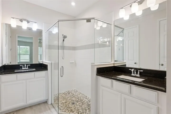 Downstairs Primary Bathroom – Bright ensuite bath features dual vanities with dark stone countertops, abundant white cabinetry, oversized mirrors, and a glass-enclosed tiled shower with a decorative pebble-style floor.