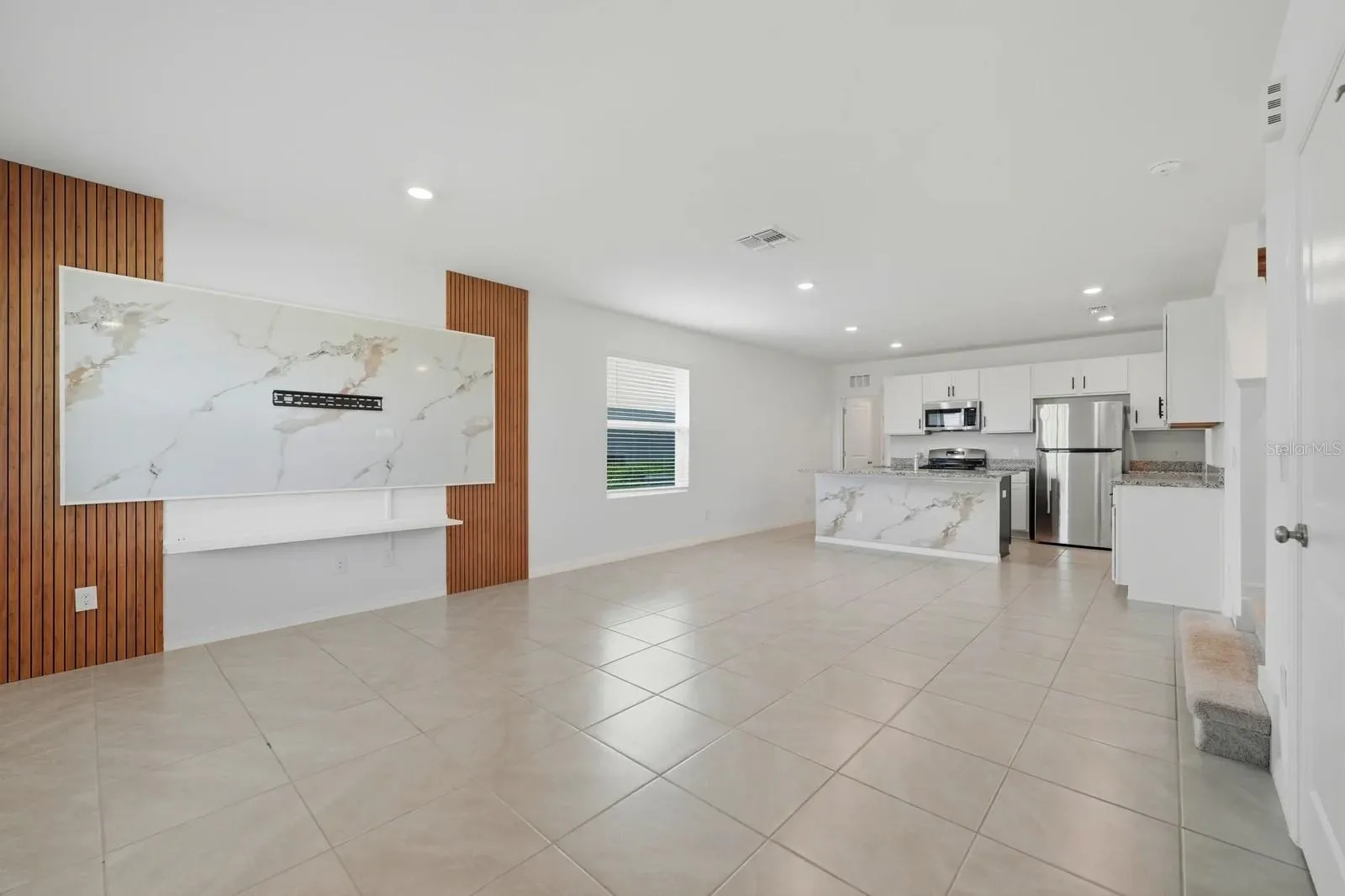 Bright open-concept living and kitchen area featuring tile flooring, stainless steel appliances, and a custom decorative TV accent wall