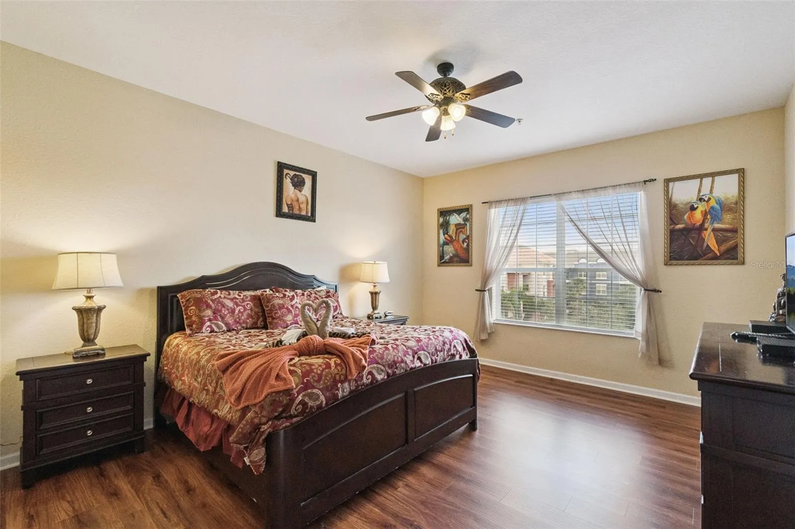 Primary Suite Layout:Spacious primary retreat displaying warm neutral tones, wood-look plank flooring, matching dark wood headboard and nightstands, ceiling fan, and a bright window.