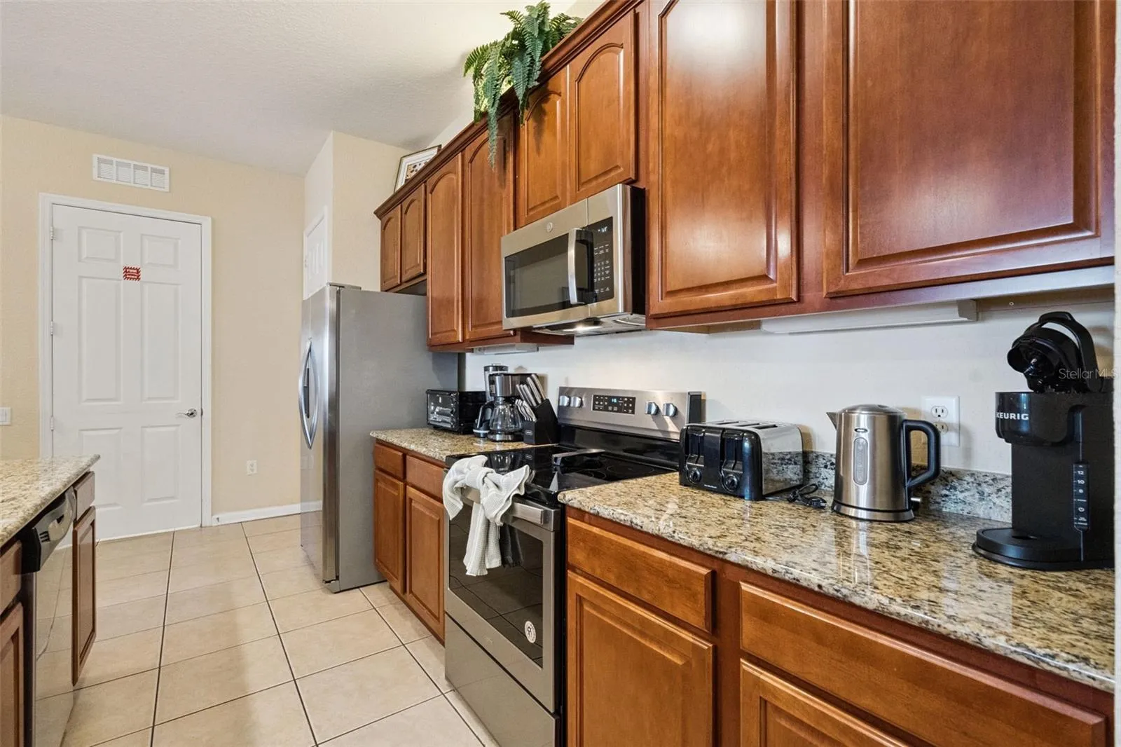 Kitchen Prep & Storage Area:Functional counter space highlighting rich wood upper and lower cabinets, granite counters, glass-top range, built-in microwave, and tile flooring.