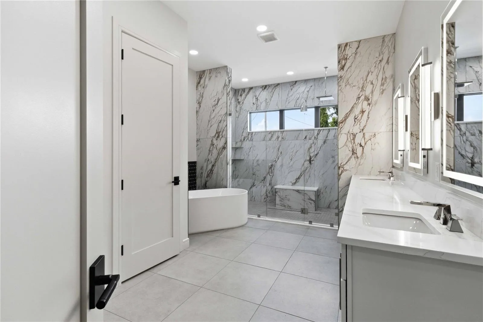 Floor-to-ceiling marble tile surrounds the oversized glass-enclosed shower, while a freestanding soaking tub, dual vanities, illuminated mirrors, contemporary fixtures, and a dedicated makeup station bring boutique-hotel luxury into everyday life