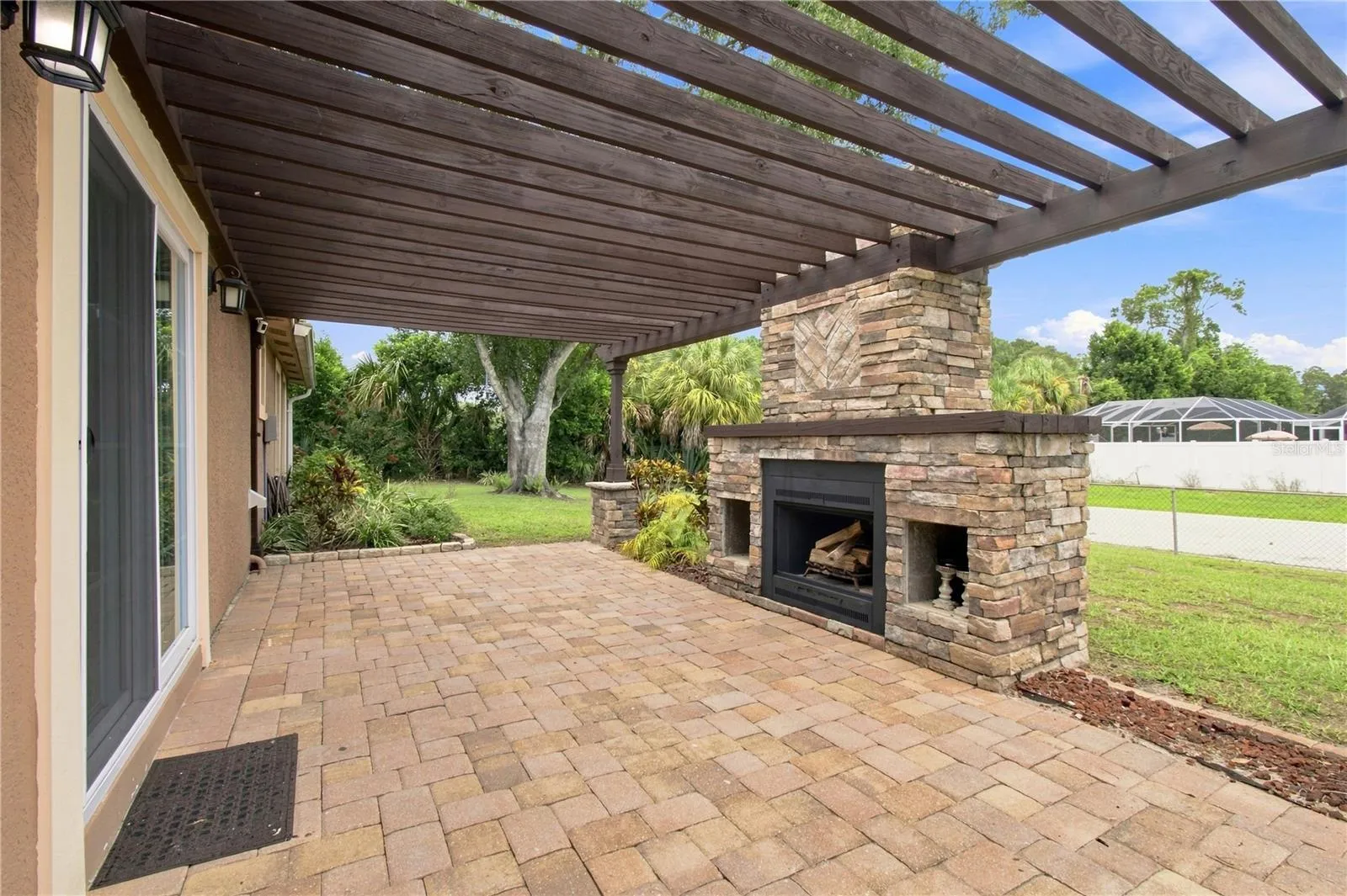 Crafted outdoor patio space featuring a large, rustic stone wood burning fireplace built under a wooden pergola and warm color pavers.