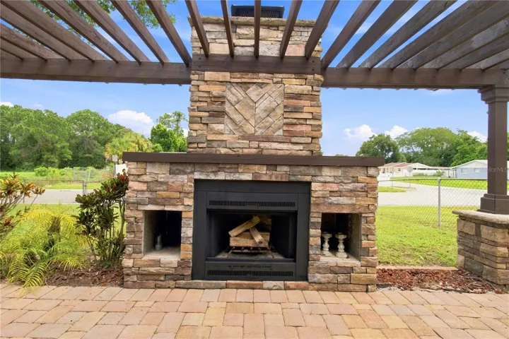 Crafted outdoor patio space featuring a large, rustic stone wood burning fireplace built under a wooden pergola and warm color pavers.