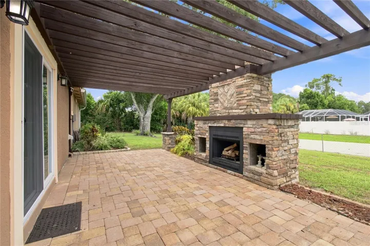 Crafted outdoor patio space featuring a large, rustic stone wood burning fireplace built under a wooden pergola and warm color pavers.