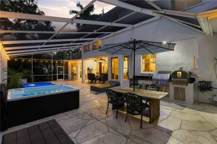 An entertainer's dream at dusk — glowing spa, umbrella-shaded dining, and a fully equipped outdoor kitchen with grill and pizza oven, all under one screened lanai.