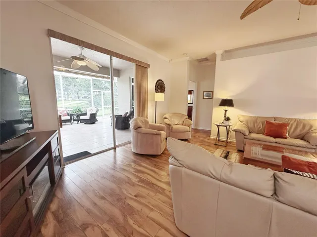 Living room with sliding glass doors onto the covered and screened in lanai with golf course view and entry to Primary Bedroom in background. Laminate flooring, crown molding, decorative shelving