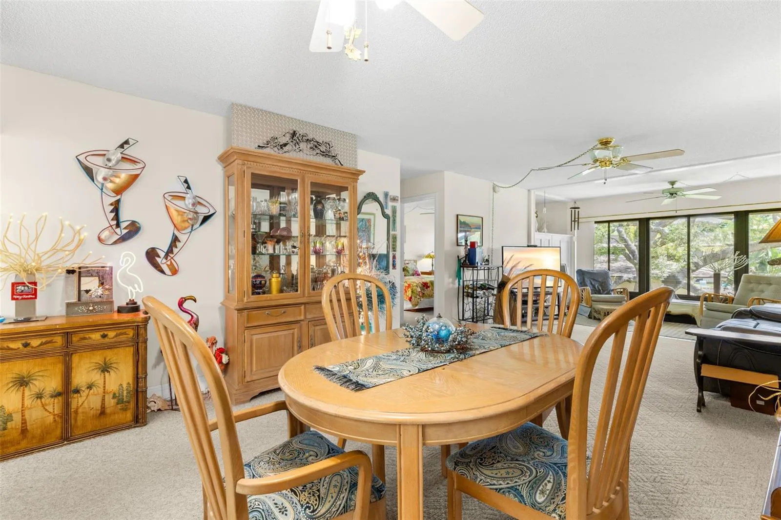 From the dining area, the sightline extends through the living room to the wall of windows at the far end. Neutral carpeting continues through both spaces, with ceiling fans overhead and the bedroom hall tucked off to the side.