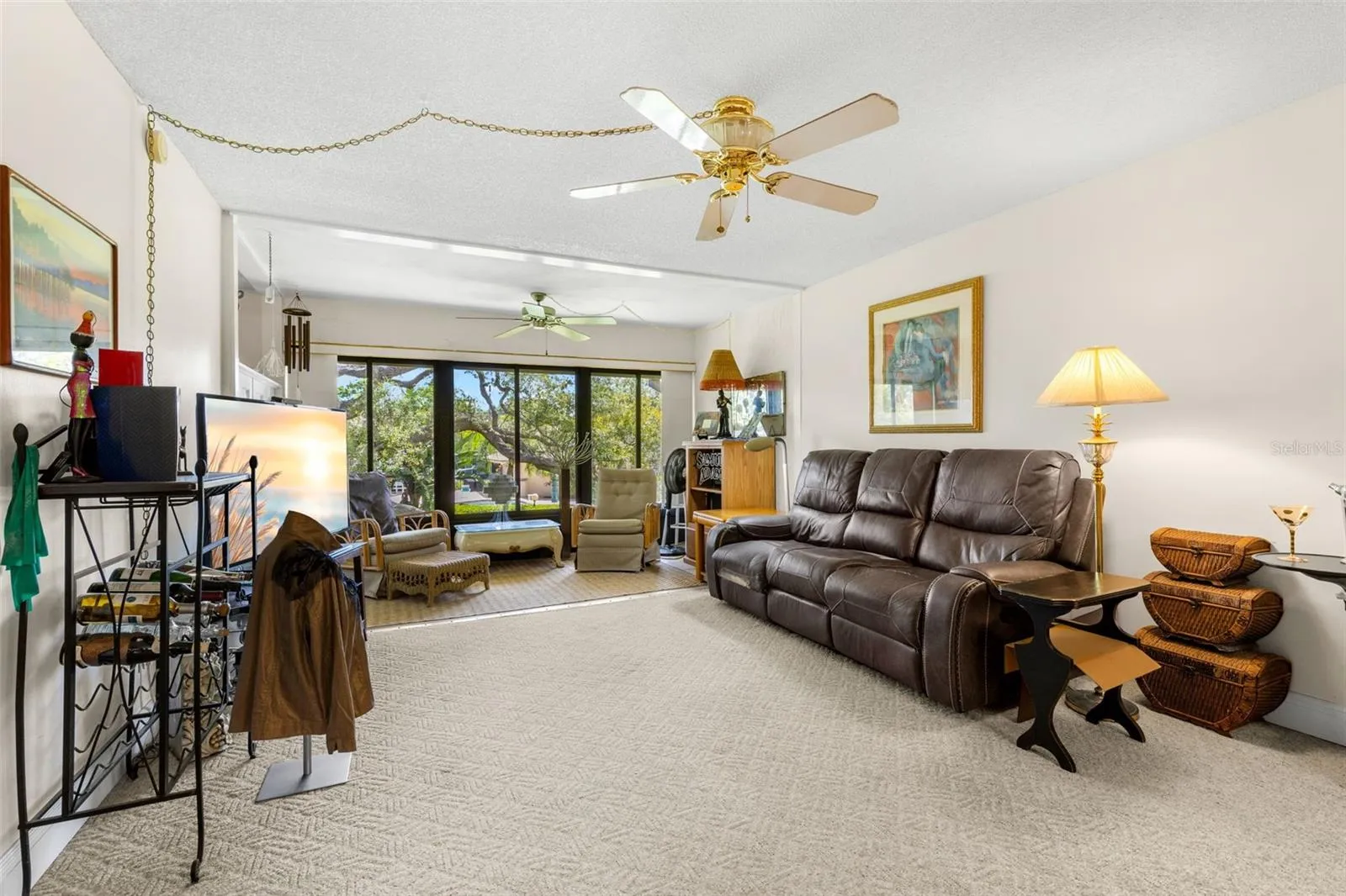 The living room extends into an enclosed Florida room, where a broad wall of windows brings in natural light and frames the mature trees outside. The extra space works comfortably as a second sitting area without interrupting the main living room.