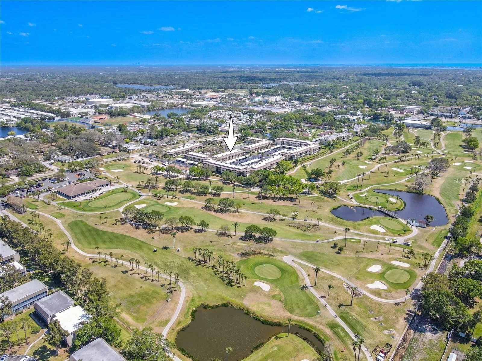Villas de Golf is arranged around landscaped grounds with the community pool and tennis courts near the center, while the neighboring golf course stretches along the east side of the property and the Gulf sits in the distance.