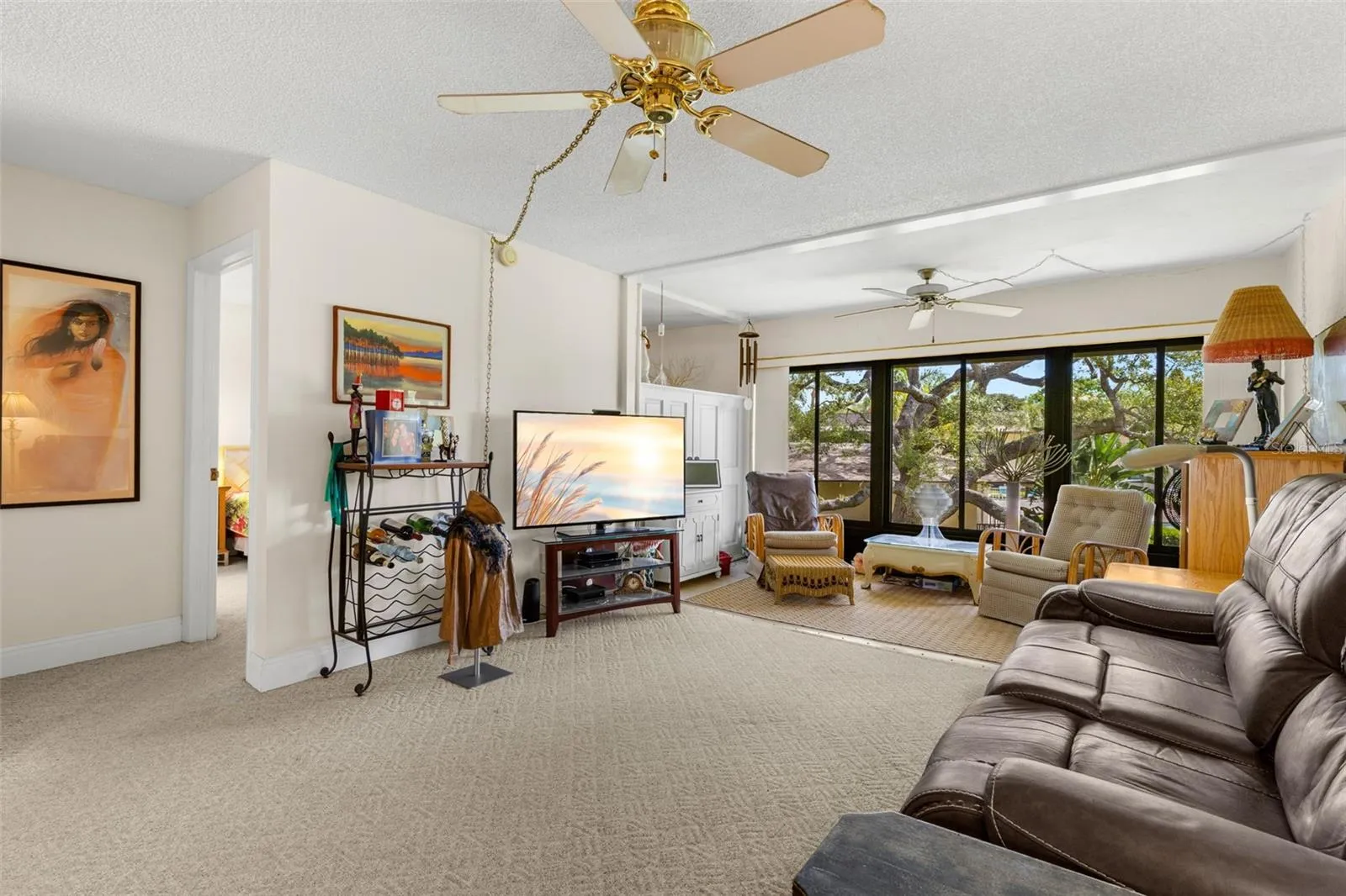 The living room opens directly into the enclosed Florida room, creating a second sitting area framed by a broad wall of windows. Neutral carpeting continues through both spaces, while ceiling fans add comfort overhead.