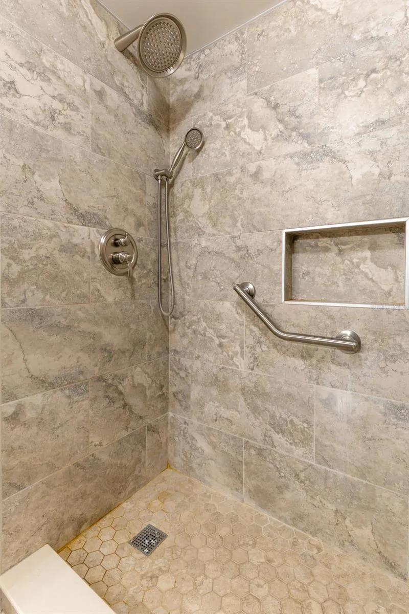 The walk-in shower is finished with large-format wall tile and a hex-pattern tile floor, with both fixed and handheld showerheads, a recessed niche and a grab bar built into the space.