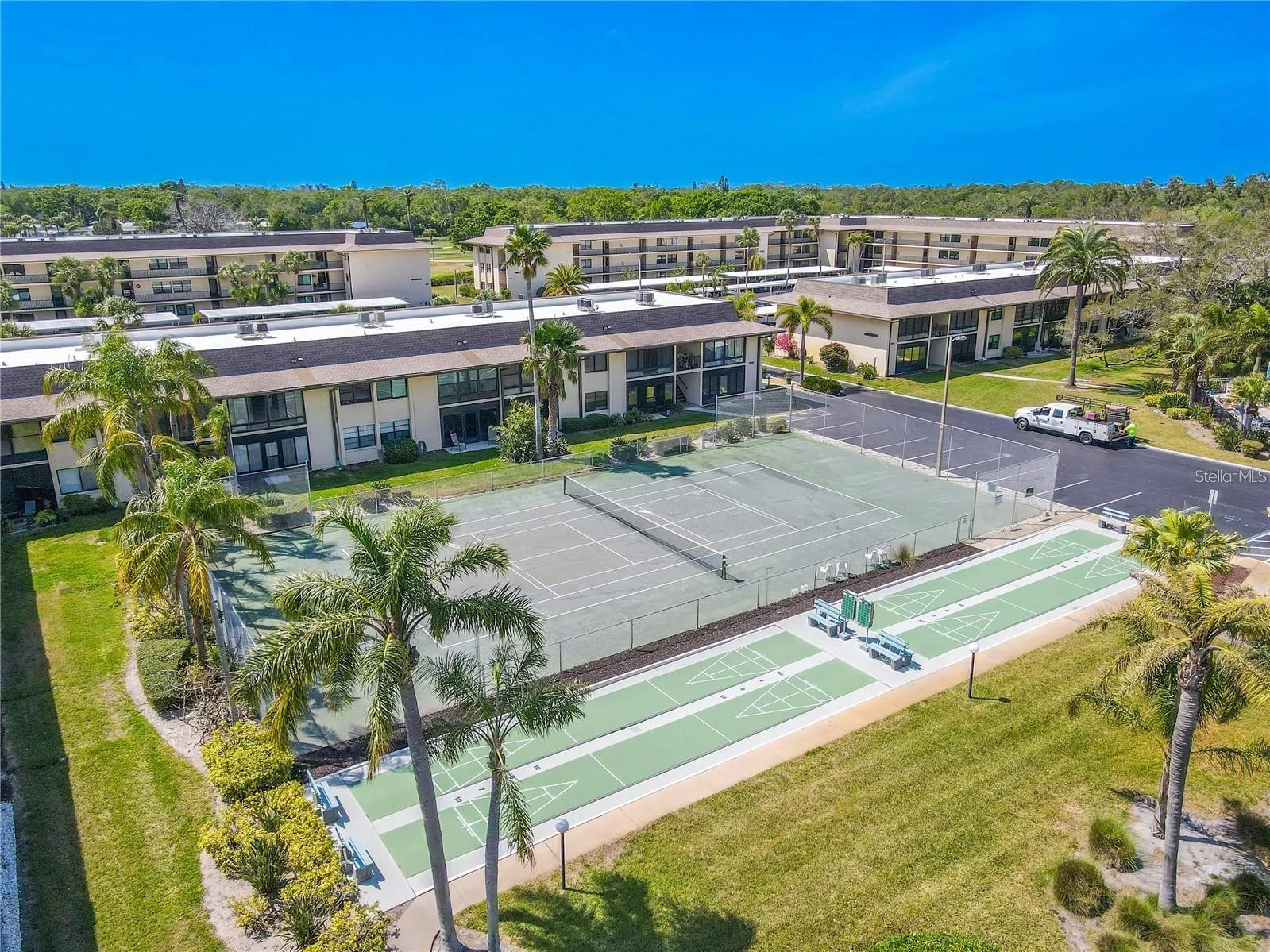 The community recreation area includes a full tennis court and multiple shuffleboard courts, with seating nearby and landscaped grounds surrounding the space.