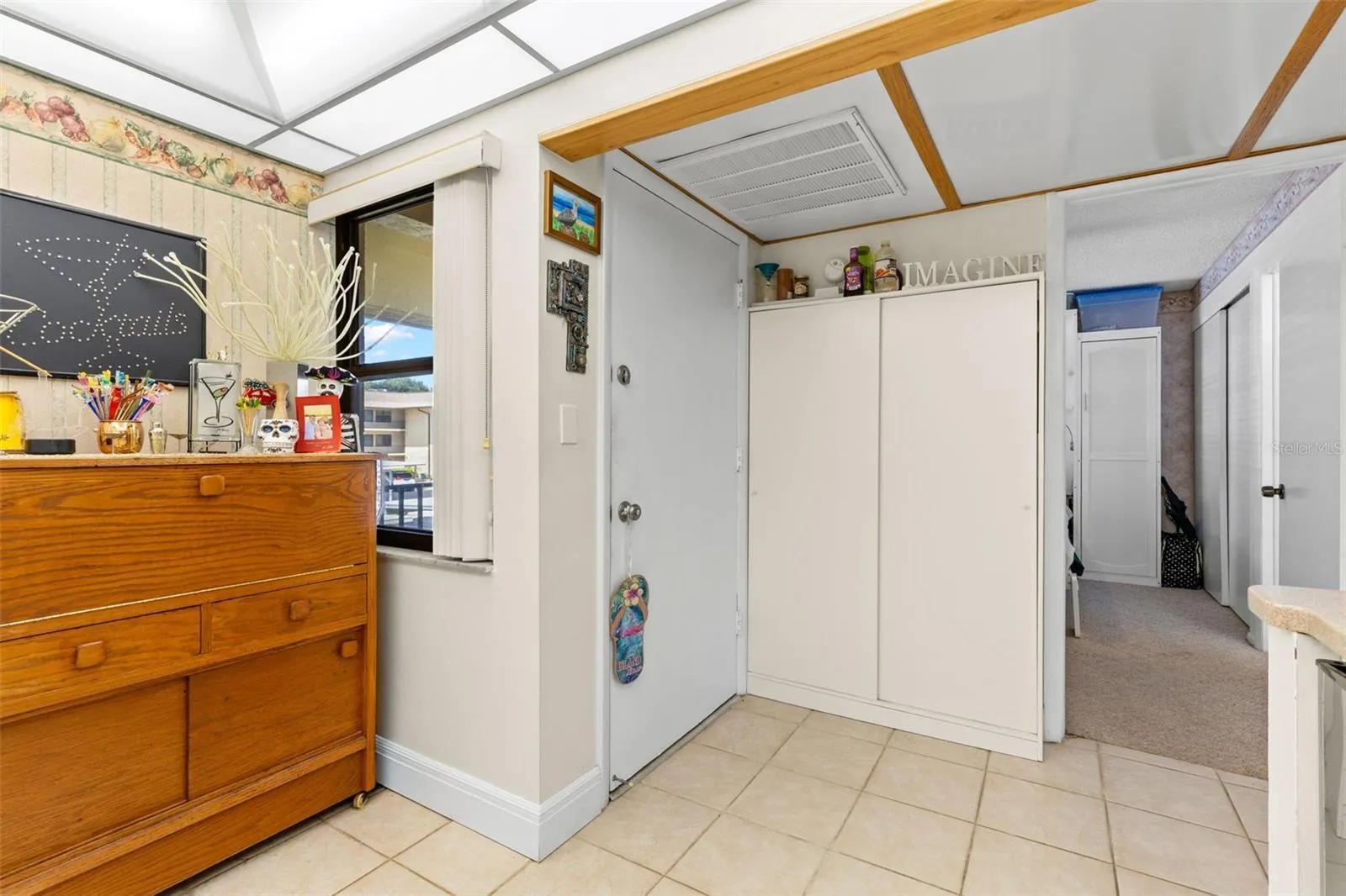 A window beside the front door brings natural light into the entry, where a tall double-door cabinet adds useful storage. Just beyond, a separate opening leads to the second bedroom and bath.