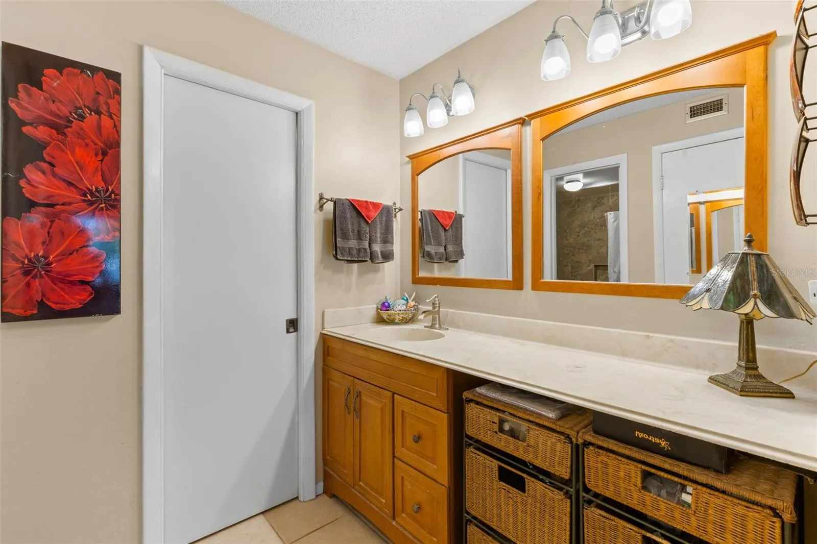 The primary bath pairs warm wood cabinetry with a long counter, framed mirrors and vanity lighting. A single sink leaves generous counter space, while the open knee space beside it provides a dedicated makeup/vanity area.