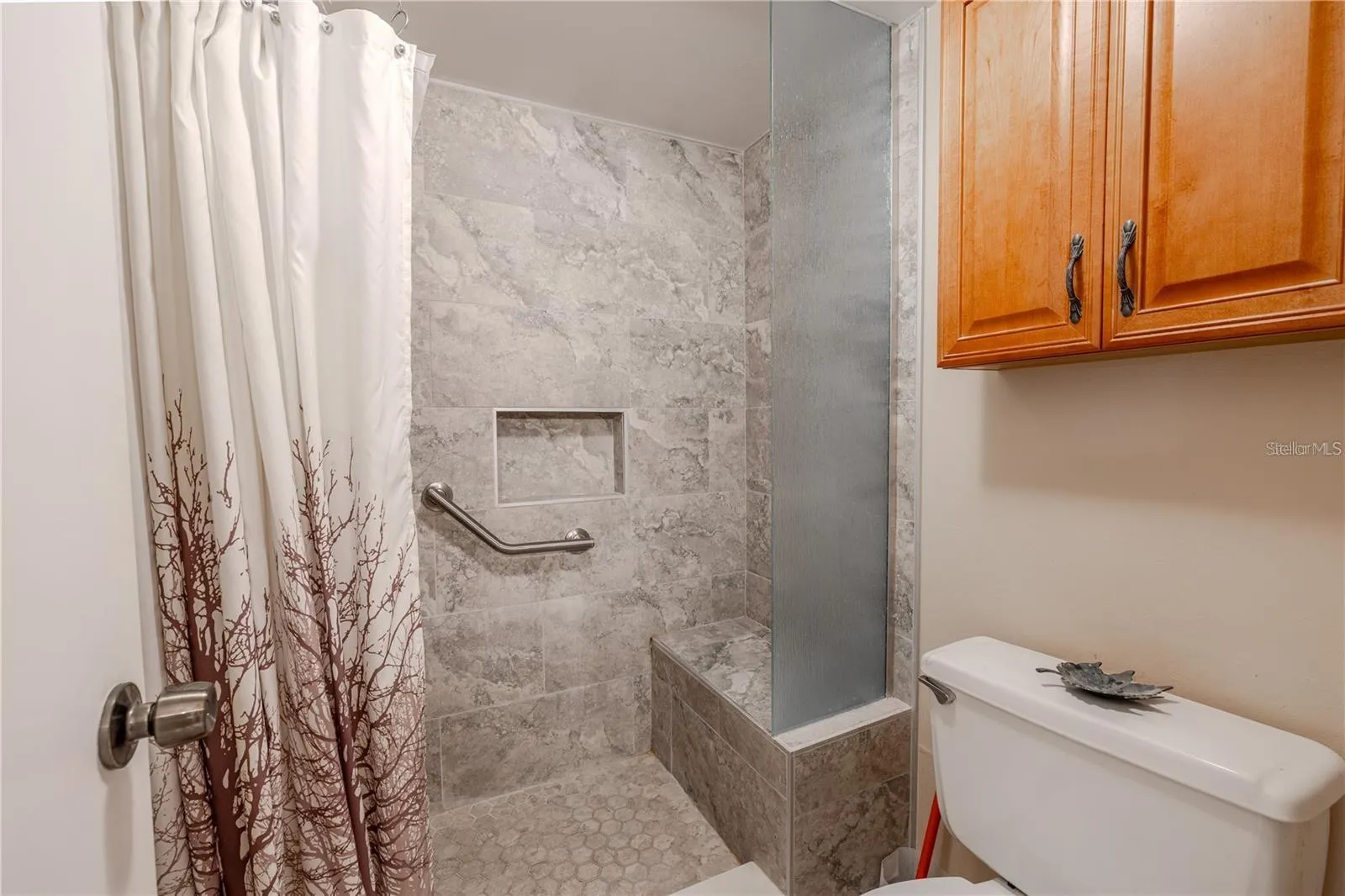 The primary bath includes a tiled walk-in shower with a built-in bench, recessed niche and grab bar. A tall textured-glass panel adds separation, while the wood cabinet above the toilet provides extra storage.