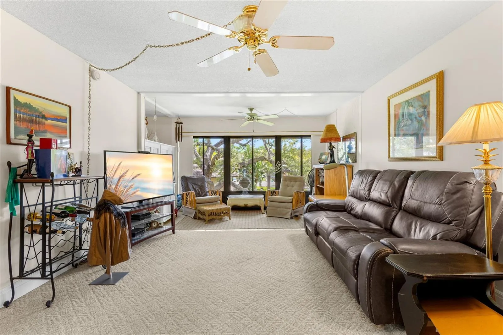 The living room stretches toward a wall of windows overlooking mature trees, with neutral carpeting and ceiling fans throughout. The length of the room also allows for a separate sitting area at the far end without crowding the main seating space.