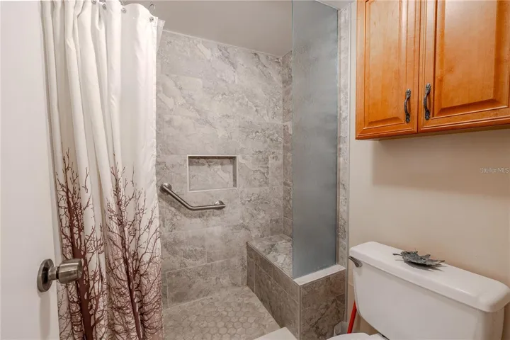 The primary bath includes a tiled walk-in shower with a built-in bench, recessed niche and grab bar. A tall textured-glass panel adds separation, while the wood cabinet above the toilet provides extra storage.