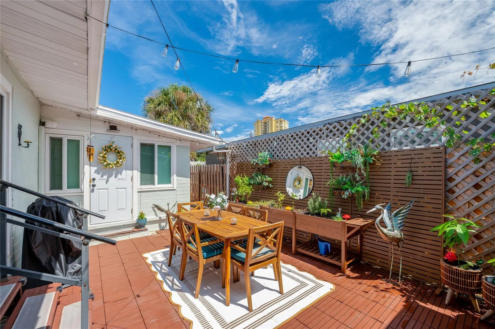 Charming outdoor wooden deck framed by lattice fencing, ambient string lights, and potted greenery. An entryway with a decorative wreath adds a welcoming touch.