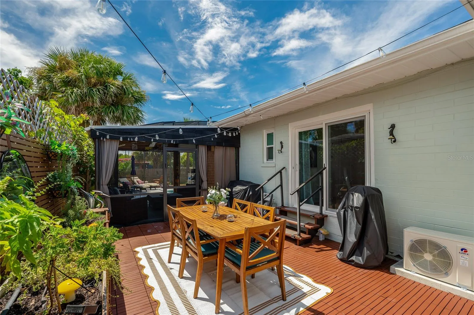 Spacious outdoor patio area with dark wooden decking, sliding glass door access, and string lights overhead. The space includes raised garden planters and a adjacent screen enclosure.