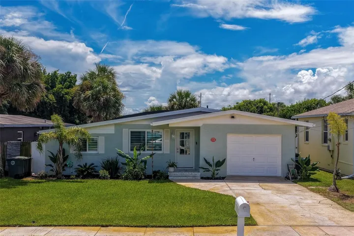 Charming single-story exterior showcasing a bright lawn, lush tropical landscaping, and a clean driveway leading to an attached garage under a vibrant blue sky.