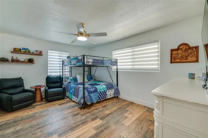 Generous bedroom design highlighting warm LVP flooring, neutral walls, two large windows with blinds, and floating display shelves.