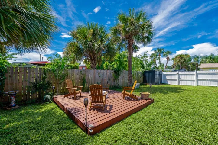 Raised wooden deck platform set within a private, fully fenced backyard. Tall palm trees overhead provide natural shade and tropical ambiance.