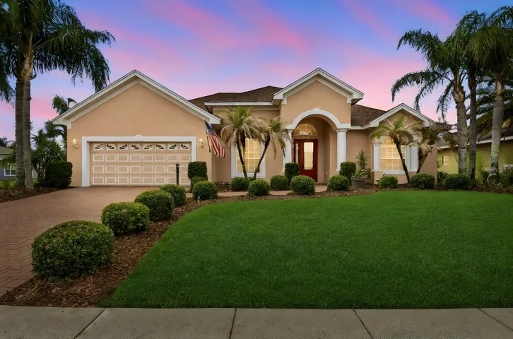 Front Exterior – Attractive single-story exterior with an arched covered entry, column accents, an attached two-car garage, brick-paver driveway, and neatly maintained landscaping framed by mature palms.