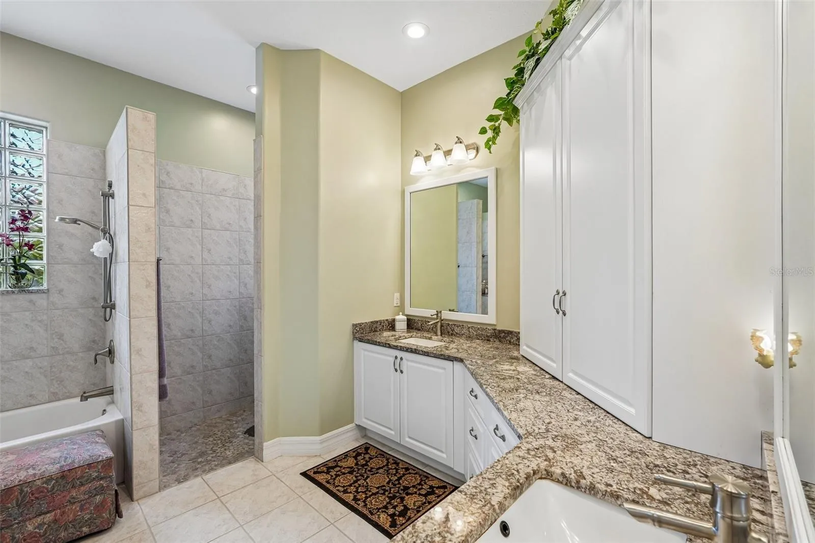 This luxurious primary en-suite bathroom boasts a expansive dual-sink vanity with granite countertops, ample storage cabinetry, a deep soaking tub and a separate tiled walk-in shower with glass block accents for privacy and light