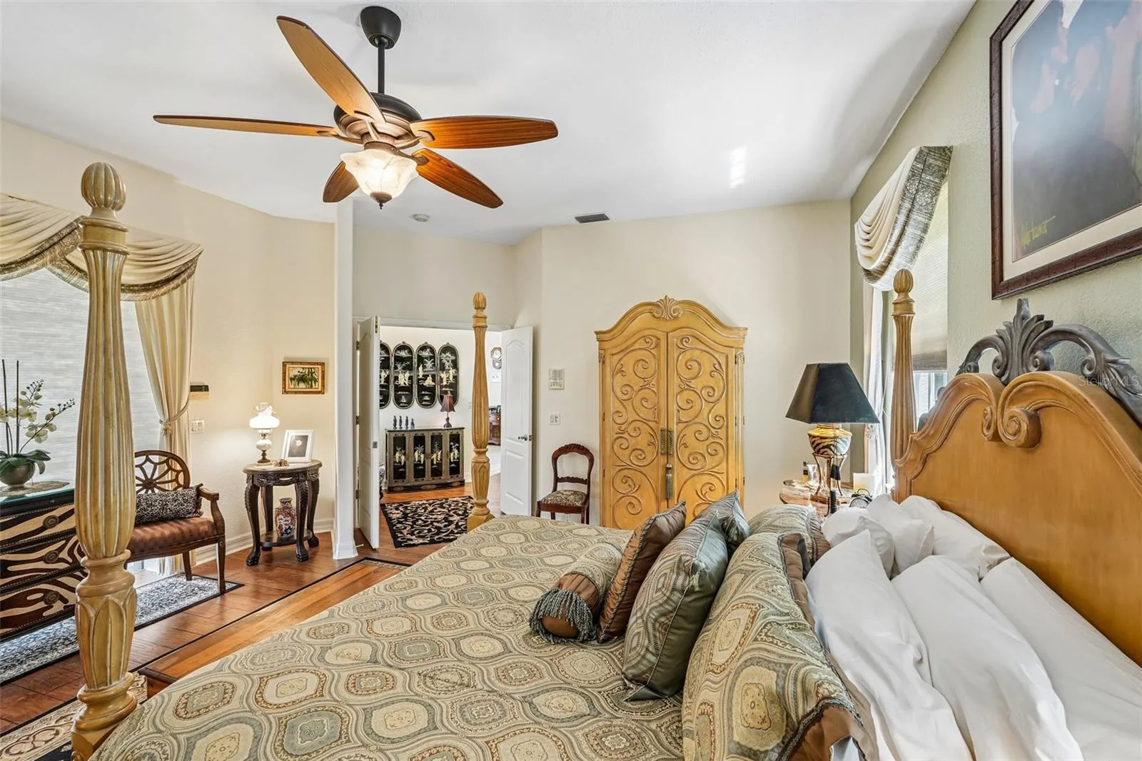 Spacious primary bedroom retreat featuring warm wood floors, elegant window treatments, a matching ceiling fan and direct access to an en-suite bathroom