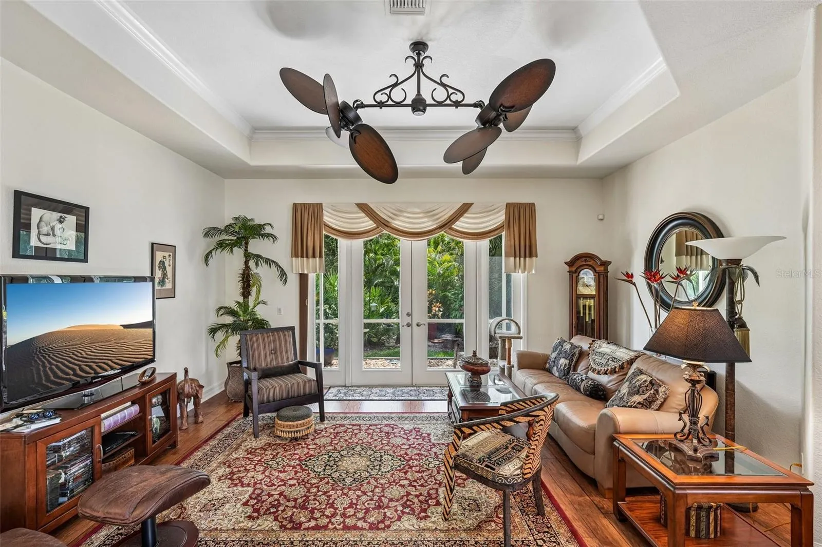 Inviting living area featuring warm wood floors, tray ceilings with crown molding, a dual-head tropical fan, and french doors offering seamless access to lush outdoor views