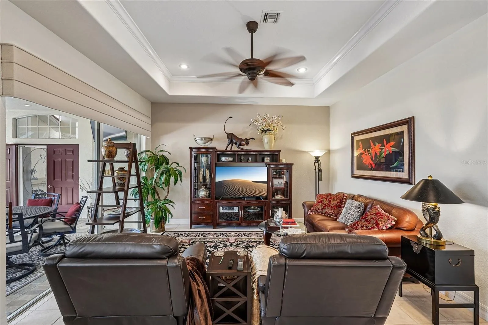 Inviting living area featuring warm wood floors, tray ceilings with crown molding, a dual-head tropical fan, and french doors offering seamless access to lush outdoor views