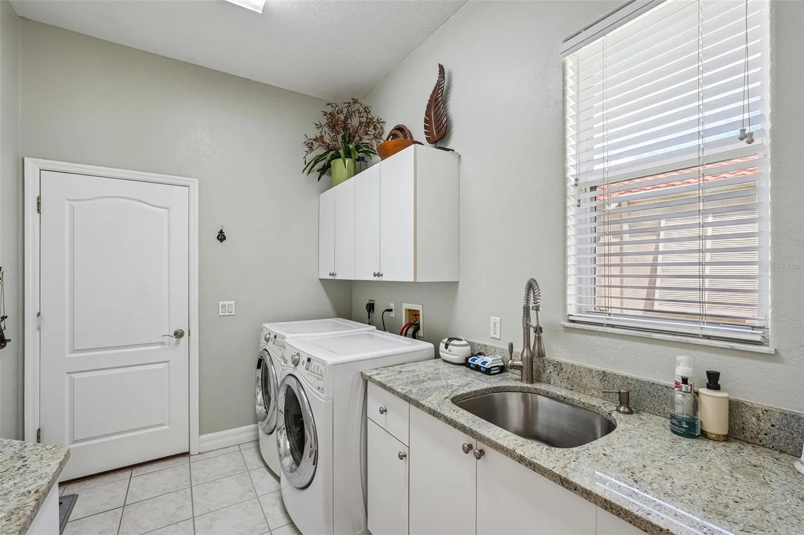 Laundry room featuring granite counter space, a built-in stainless steel utility sink, front-loading washer and dryer, tile flooring and upper and lower storage cabinets