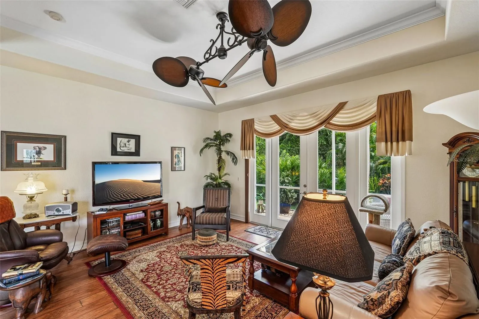 Inviting living area featuring warm wood floors, tray ceilings with crown molding, a dual-head tropical fan, and french doors offering seamless access to lush outdoor views