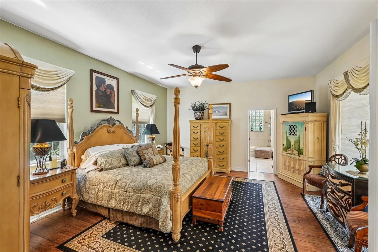 Spacious primary bedroom retreat featuring warm wood floors, elegant window treatments, a matching ceiling fan and direct access to an en-suite bathroom