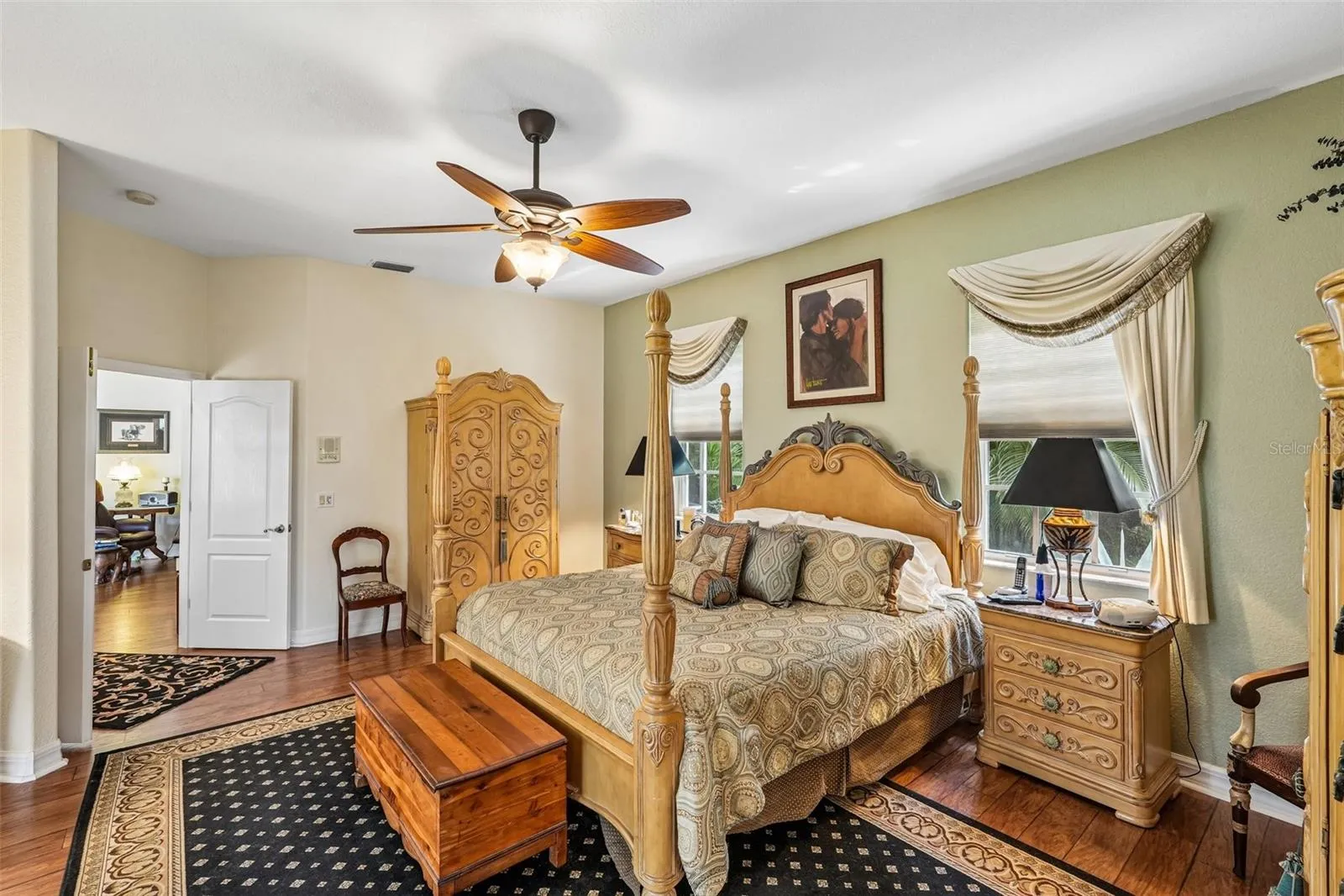Spacious primary bedroom retreat featuring warm wood floors, elegant window treatments, a matching ceiling fan and direct access to an en-suite bathroom