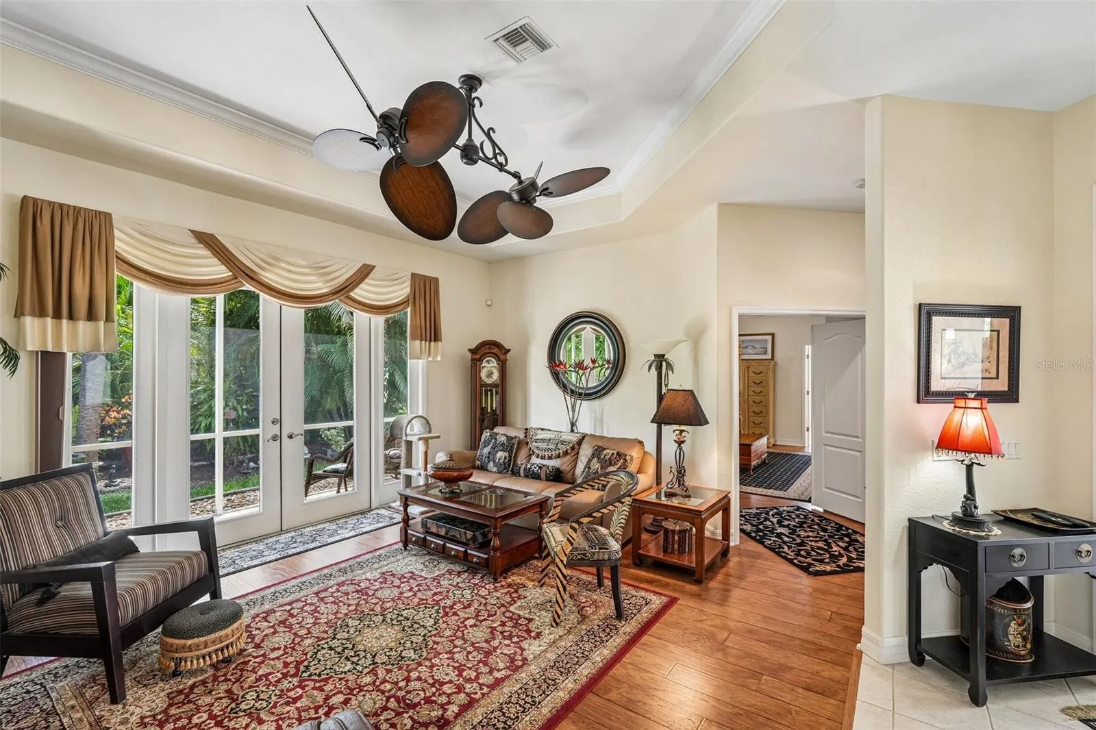 Inviting living area featuring warm wood floors, tray ceilings with crown molding, a dual-head tropical fan, and french doors offering seamless access to lush outdoor views