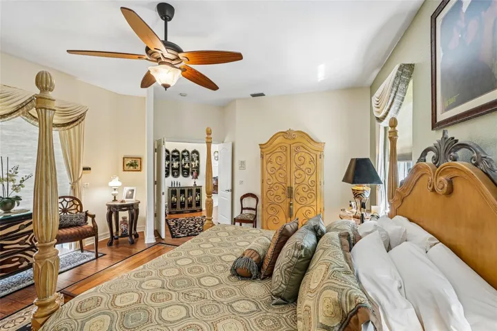 Spacious primary bedroom retreat featuring warm wood floors, elegant window treatments, a matching ceiling fan and direct access to an en-suite bathroom