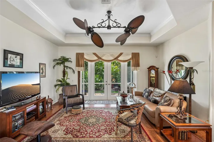 Inviting living area featuring warm wood floors, tray ceilings with crown molding, a dual-head tropical fan, and french doors offering seamless access to lush outdoor views