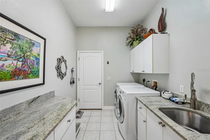 Laundry room featuring granite counter space, a built-in stainless steel utility sink, front-loading washer and dryer, tile flooring and upper and lower storage cabinets
