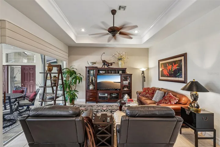 Inviting living area featuring warm wood floors, tray ceilings with crown molding, a dual-head tropical fan, and french doors offering seamless access to lush outdoor views