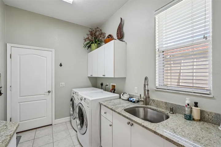 Laundry room featuring granite counter space, a built-in stainless steel utility sink, front-loading washer and dryer, tile flooring and upper and lower storage cabinets